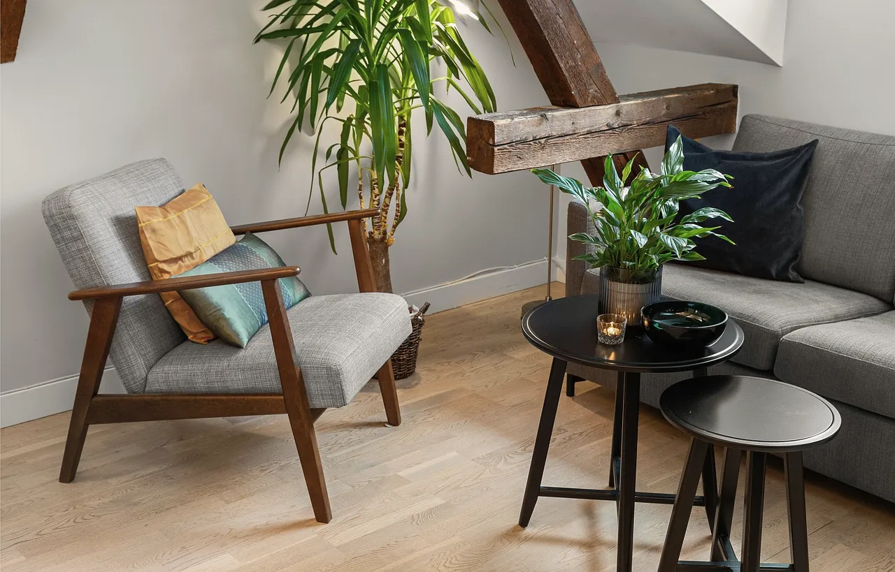 A quiet seating corner: an armchair with two cushions, a tall plant, two small round tables and the end of a sofa.